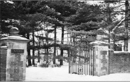 黑白照片，一个锻铁大门，两侧的砖柱与砖墙相连. Trees fill the space behind the gate, which is open.